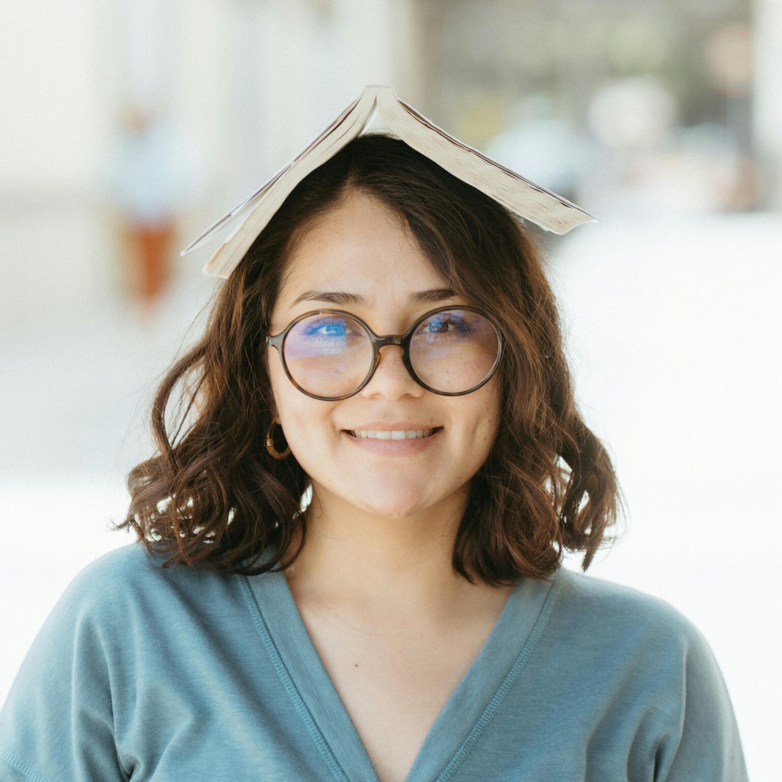 Cheerful woman wears a book on her head while smiling outside, enjoying a sunny day.