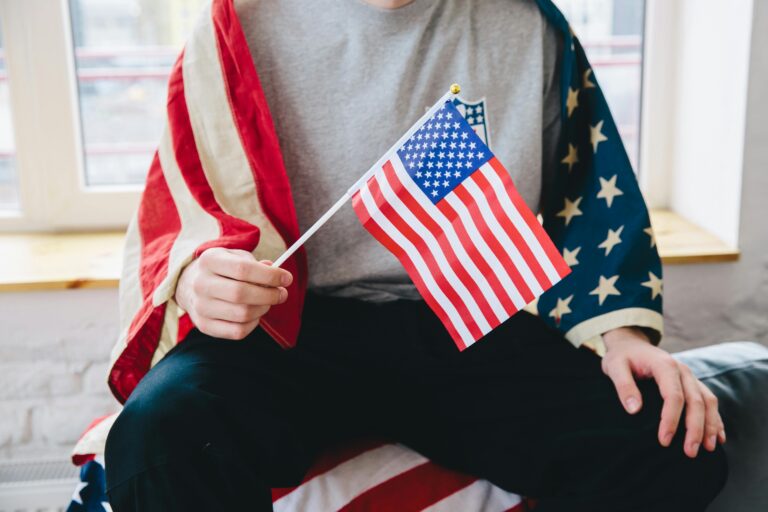 A person sitting indoors, holding a small American flag, draped in a larger flag, celebrating Independence Day.